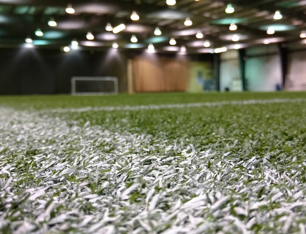 An indoor sports facility with artificial turf and overhead lighting. The focus is on the white boundary line on the field, and a soccer goal is visible in the distance. The lighting creates a well-illuminated environment with shadows cast on the turf.