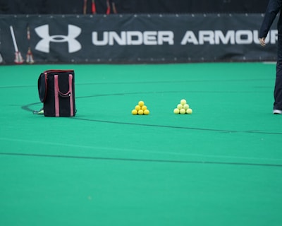 A sports field features a black and pink sports bag, two pyramids of yellow balls, and a portion of a person in dark athletic gear. The background displays a banner with the Under Armour logo.