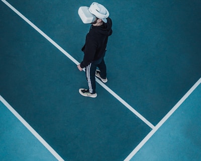 A person wearing a virtual reality headset stands on a large, empty sport court with distinct white lines on the floor. The dark clothing contrasts with the bright headset.