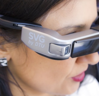 A person is wearing a pair of advanced augmented reality glasses with the label 'SVG by DTU' on the side, indicating a technological product. The glasses have a sleek, modern design and cover the eyes of the individual. The person has dark hair and is seen in a close-up view, focusing primarily on the device.