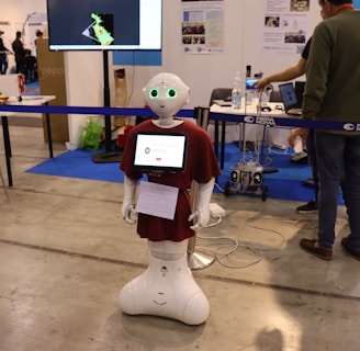 A humanoid robot with green-lit eyes and a screen on its chest displaying a logo stands in an exhibition space. It is surrounded by various pieces of equipment and people interacting with different technology displays. The setting appears to be industrial with smooth flooring, tables, chairs, and informational posters on the wall.