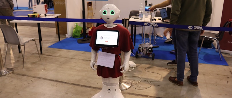 A humanoid robot with green-lit eyes and a screen on its chest displaying a logo stands in an exhibition space. It is surrounded by various pieces of equipment and people interacting with different technology displays. The setting appears to be industrial with smooth flooring, tables, chairs, and informational posters on the wall.