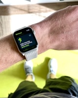 A close-up view of a person's wrist wearing a smartwatch displaying a workout mode for High Intensity Interval Training. The background includes a bright green exercise mat on a wooden floor. The person is wearing white athletic shoes and dark shorts.