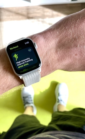 A close-up view of a person's wrist wearing a smartwatch displaying a workout mode for High Intensity Interval Training. The background includes a bright green exercise mat on a wooden floor. The person is wearing white athletic shoes and dark shorts.