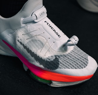 A close-up view of a modern athletic shoe featuring a sleek white and gray design with a prominent locking mechanism labeled 'LOCK' and text 'FLYEASE'. The shoe has colorful accents including a bright pink and orange sole.