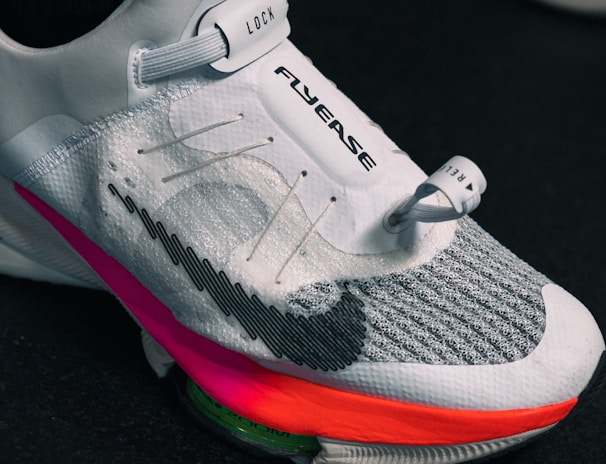 A close-up view of a modern athletic shoe featuring a sleek white and gray design with a prominent locking mechanism labeled 'LOCK' and text 'FLYEASE'. The shoe has colorful accents including a bright pink and orange sole.