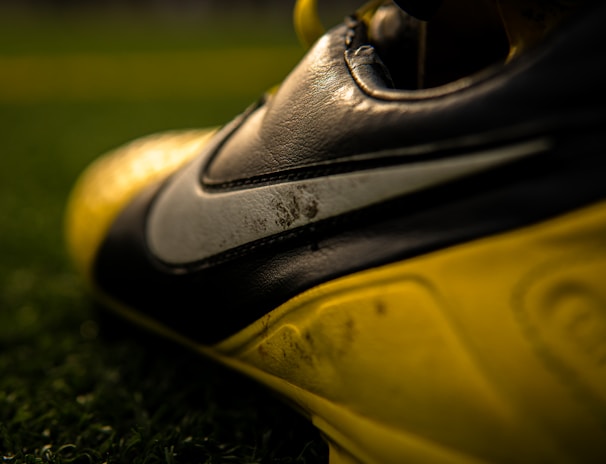 A close-up view of a football or soccer shoe with a distinctive swoosh logo in focus, featuring scuffed and worn textures on the leather surface. The shoe is predominantly black and yellow, with rich detailing captured under gleaming light. The background includes blurred green artificial turf, suggesting a sports field setting.