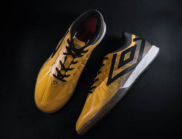 A pair of yellow and black sports shoes displayed against a dark background. The shoes feature a bold geometric pattern and are designed for athletic performance.