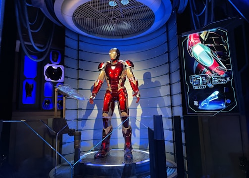 A high-tech suit displayed in a futuristic setting, illuminated by focused lighting. The surroundings include digital screens showing technical data and designs related to the suit. The environment appears to be a lab or exhibit with metallic and technological elements.