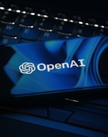 A smartphone displaying the OpenAI logo rests on a laptop keyboard. The screen features a blue abstract design, and the keyboard is visible beneath with dimly lit keys.