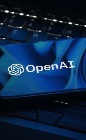 A smartphone displaying the OpenAI logo rests on a laptop keyboard. The screen features a blue abstract design, and the keyboard is visible beneath with dimly lit keys.