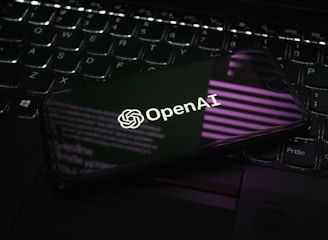 A smartphone displaying the OpenAI logo is resting on a laptop keyboard. The phone screen reflects purple and white light patterns, adding a modern and tech-focused ambiance.