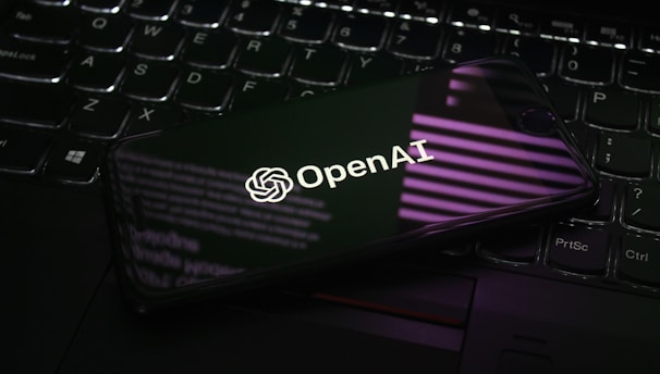 A smartphone displaying the OpenAI logo is resting on a laptop keyboard. The phone screen reflects purple and white light patterns, adding a modern and tech-focused ambiance.