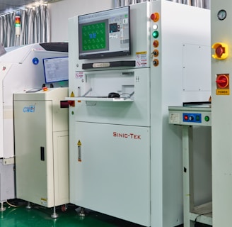 A laboratory setting with several advanced machines, including a prominent white machine labeled 'SINIC-TEK' featuring a large monitor on top displaying technical graphics. Beside it, another machine displays the label 'GWEI' with a smaller screen. The setting has a high-tech and industrial appearance with a polished green floor and metallic curtains.