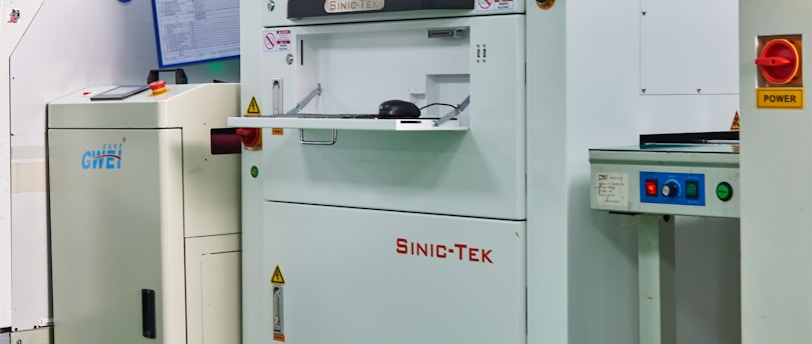 A laboratory setting with several advanced machines, including a prominent white machine labeled 'SINIC-TEK' featuring a large monitor on top displaying technical graphics. Beside it, another machine displays the label 'GWEI' with a smaller screen. The setting has a high-tech and industrial appearance with a polished green floor and metallic curtains.