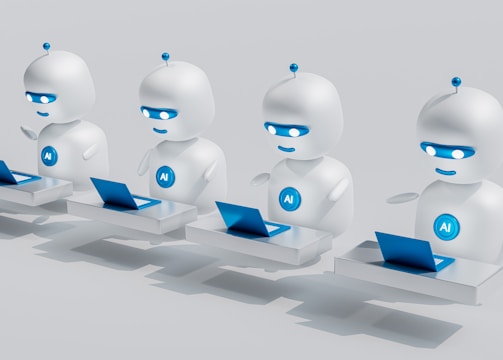 Four small, white robot figures are sitting in a row, each with a laptop in front of them. They have round bodies and blue accents, with 'AI' displayed on their chests. The background is minimal and light gray, emphasizing the robots and their computers.