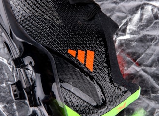 A close-up view of a sleek black sports shoe with a textured surface and an orange logo. The sole has a shiny finish with molded studs, and there is a neon green accent at the bottom.