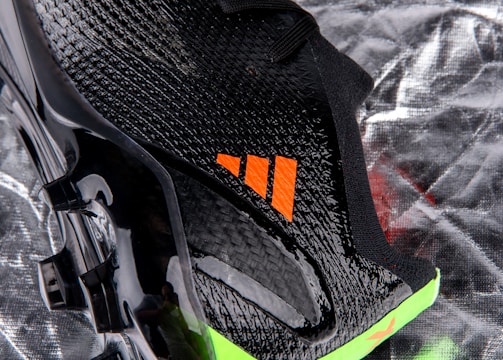 A close-up view of a sleek black sports shoe with a textured surface and an orange logo. The sole has a shiny finish with molded studs, and there is a neon green accent at the bottom.